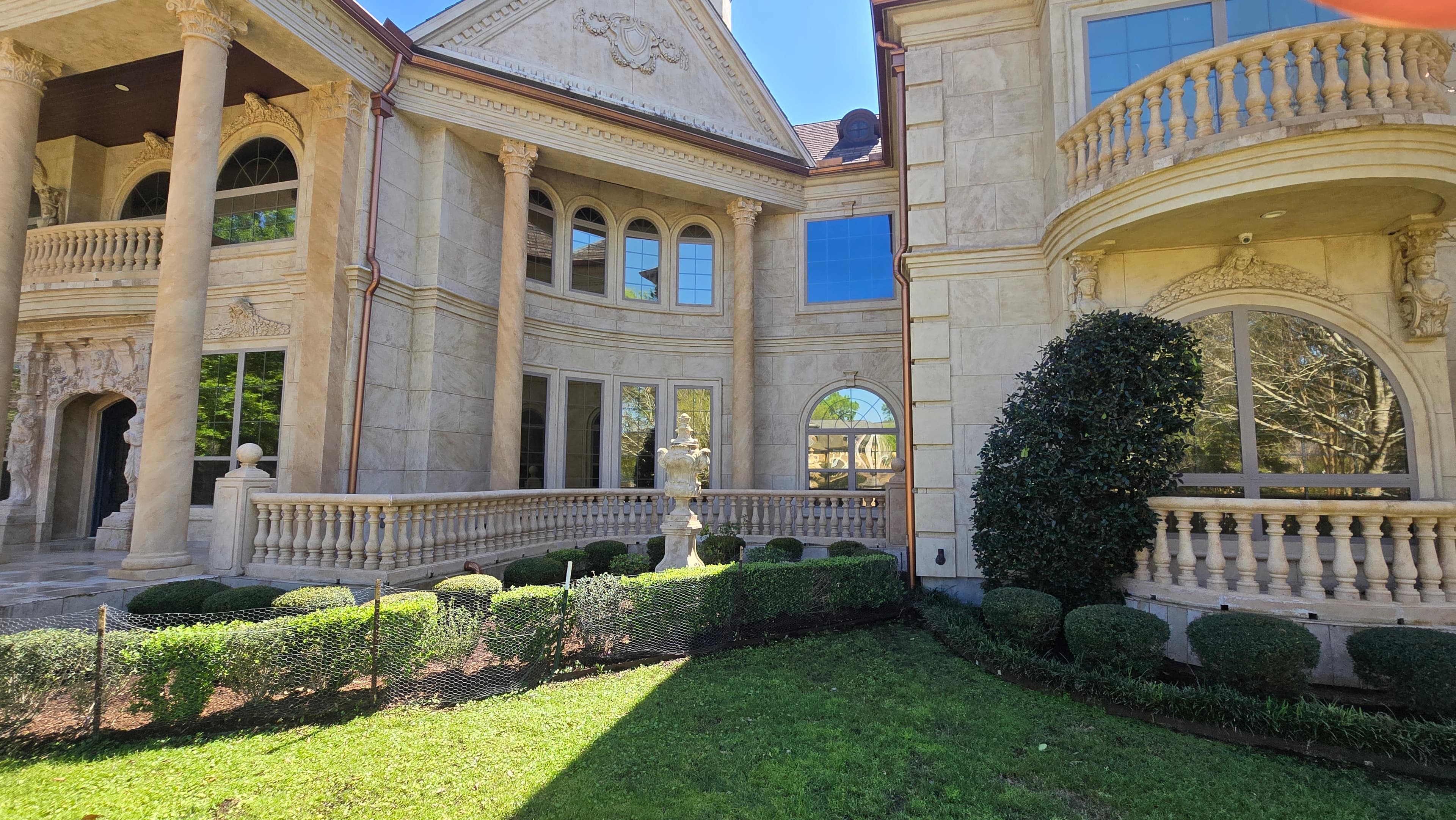 Ornate stone mansion featuring classical columns, arched windows, and manicured hedges in a sunny courtyard.