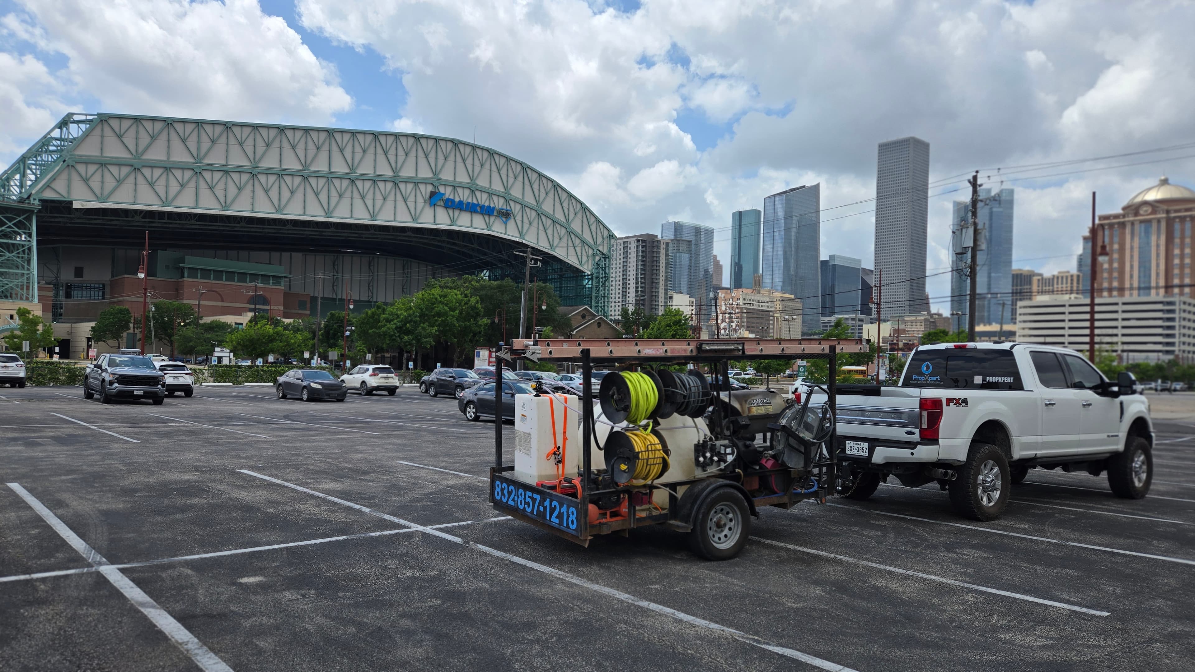 White truck towing pressure washing trailer in parking lot near Daikin Park and city skyline.
