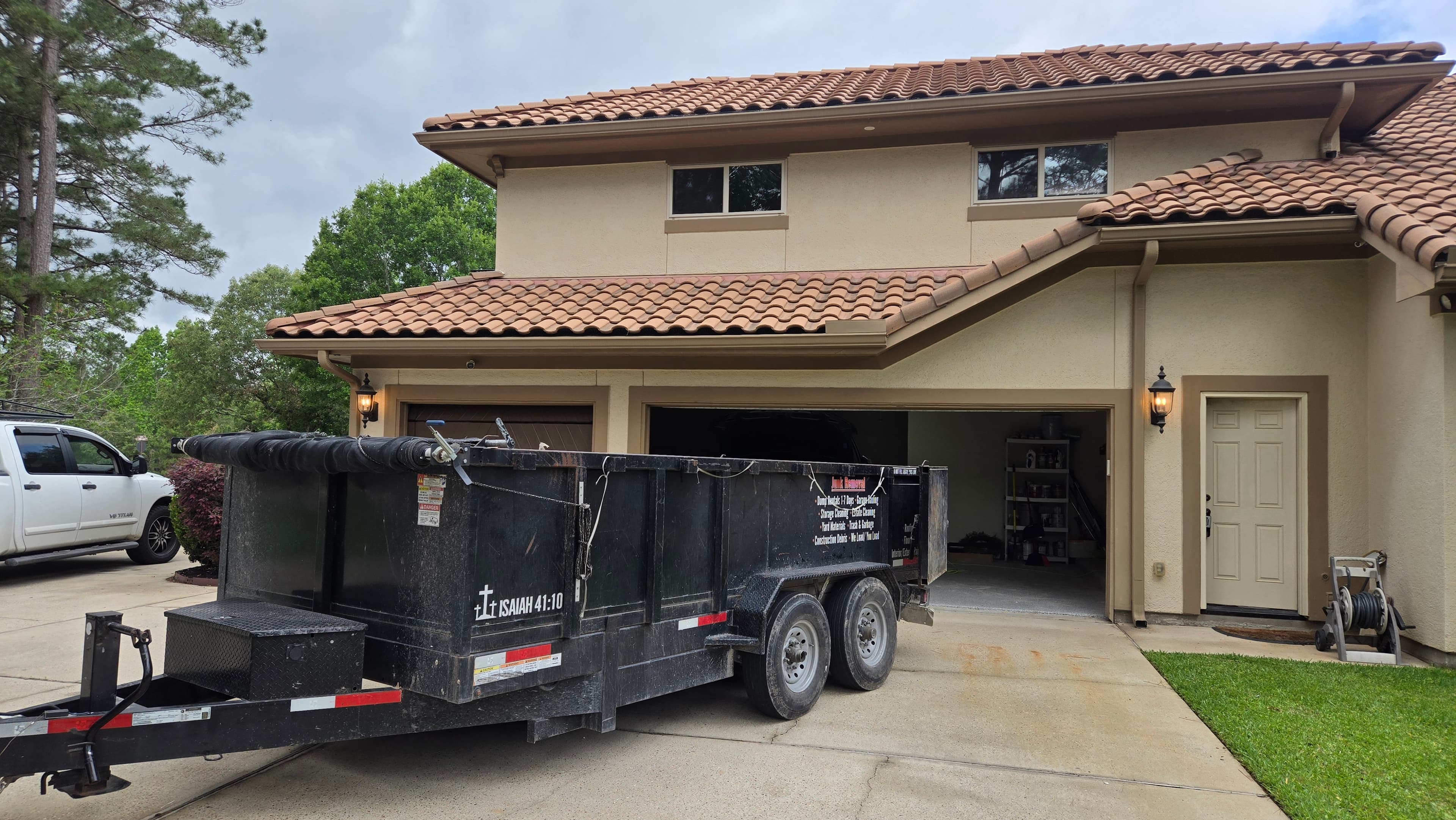 Black junk removal trailer parked in the driveway of a tan house with tile roof.