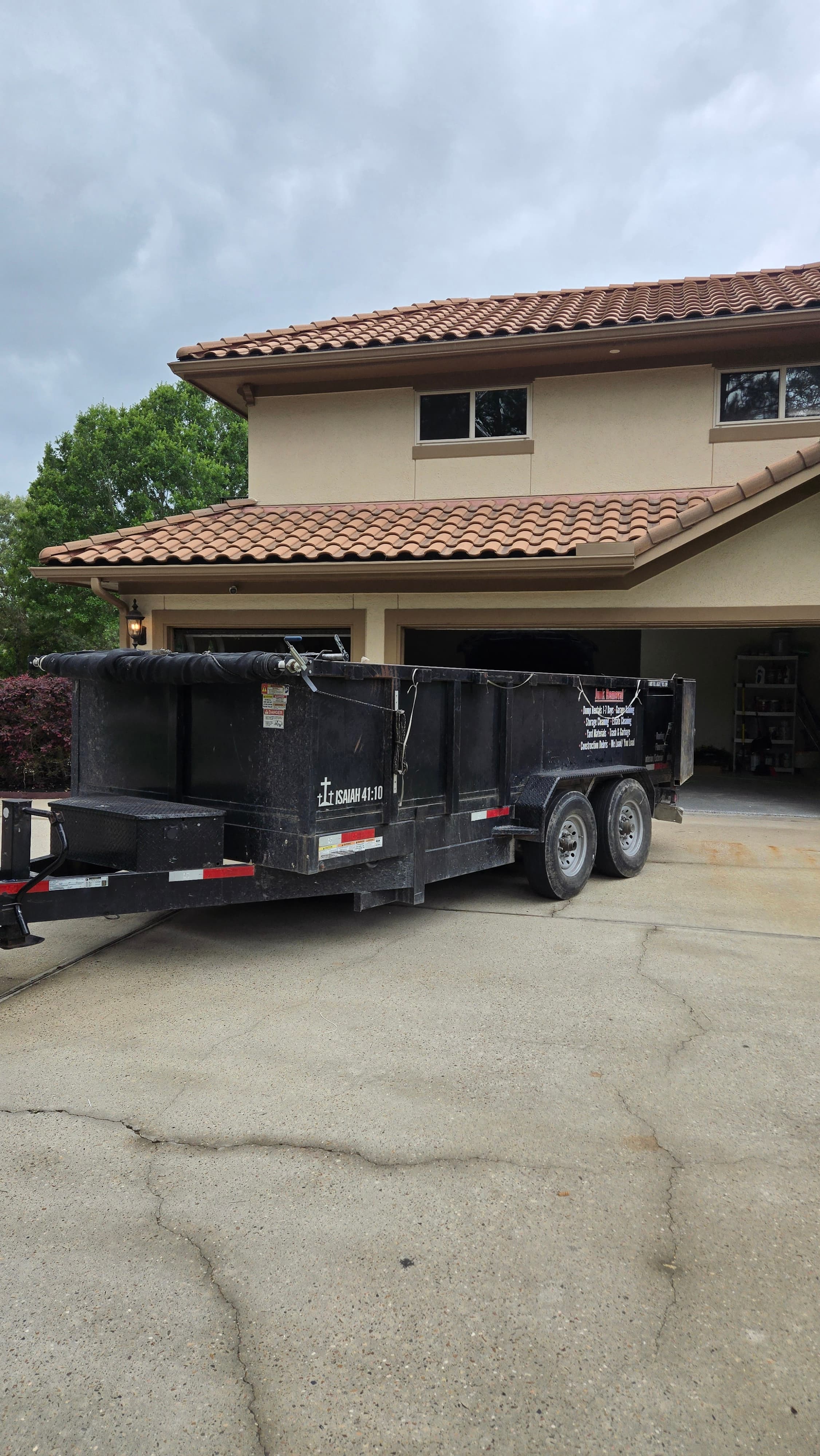 Black dump trailer parked on a concrete driveway beside a beige house with terracotta roof.