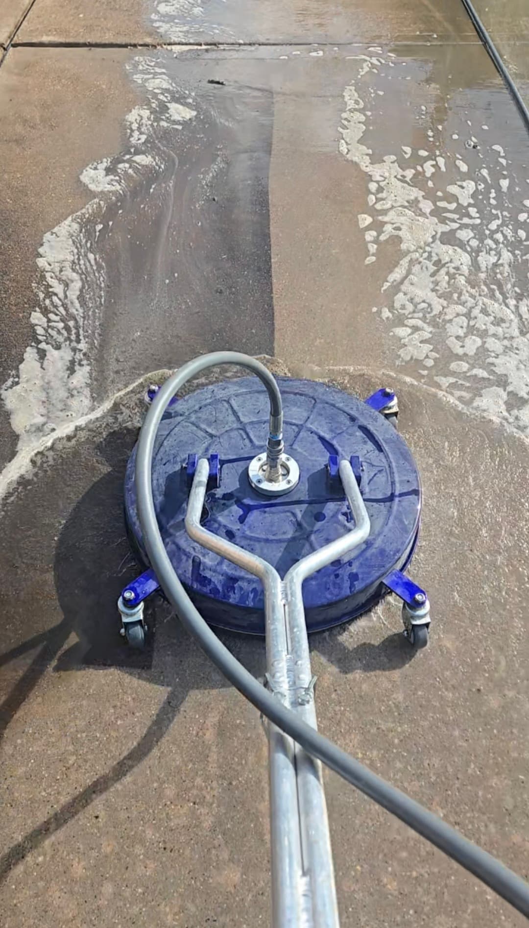 Blue circular surface cleaner pressure washing a concrete driveway, revealing a clean path through foam.