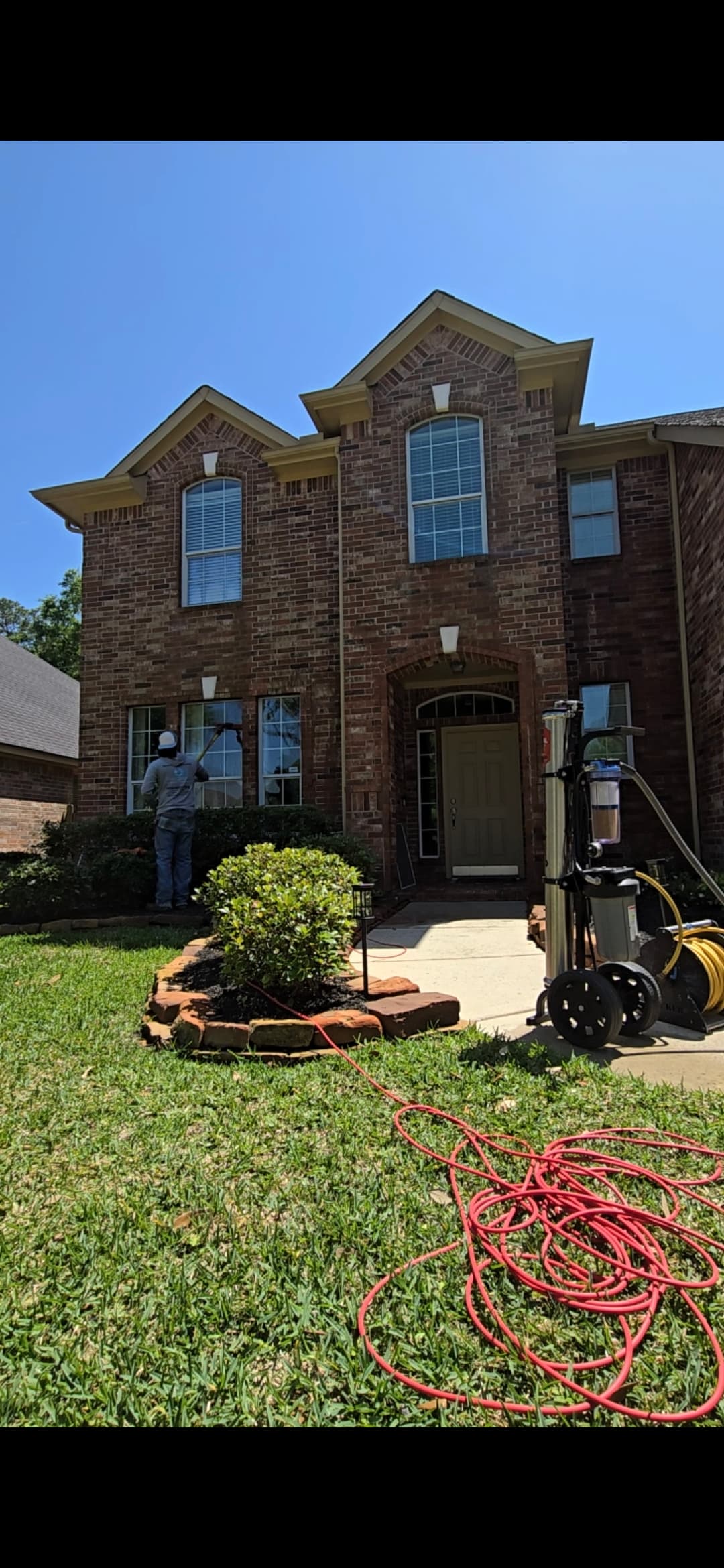 Professional window cleaner using a water-fed pole to wash windows of a brick house.