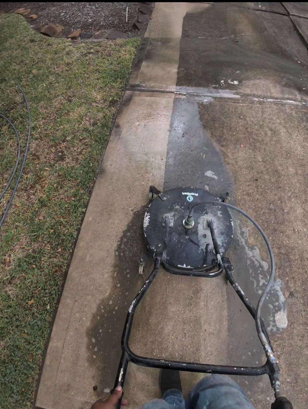A circular surface cleaner pressure washing a driveway, revealing a clean path on dirty concrete.